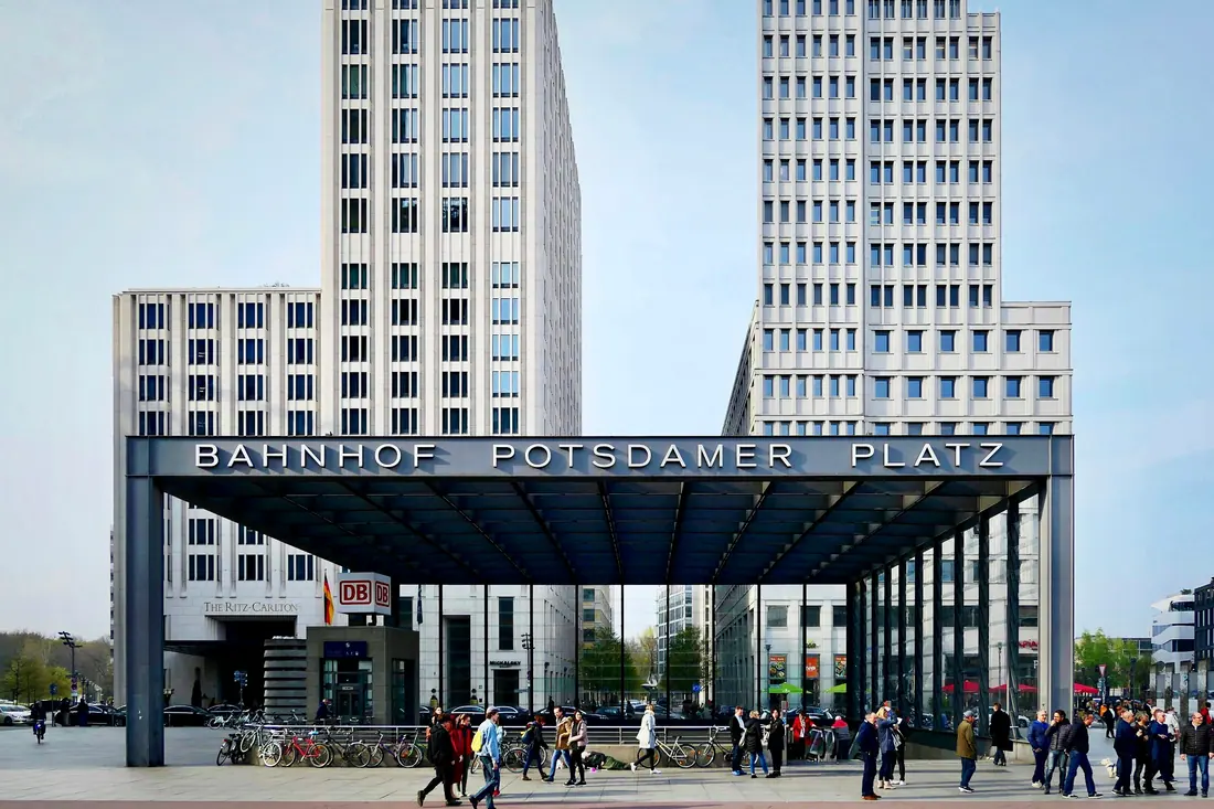 The Bahnhof Potsdamer Platz entrance canopy with tall towers rising behind it and people crossing the square